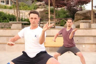 Two men practicing a taijiquan stance outdoors, one positioned in front of the other