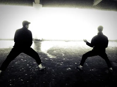 Two people facing each other in a low taijiquan stance, silhouetted against bright light