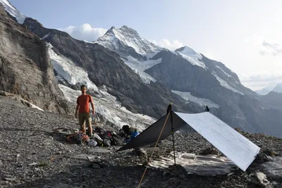 A mountaineer at a tarp bivouac on a rocky moraine below a glaciated alpine peak