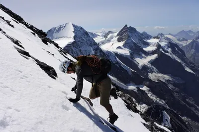 A climber in a helmet ascending a steep snow slope with alpine peaks behind