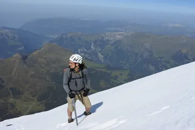 A mountaineer with an ice axe standing on a snow slope high above green alpine valleys
