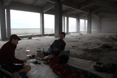 Two men resting with a tea set on the floor of an unfinished concrete building